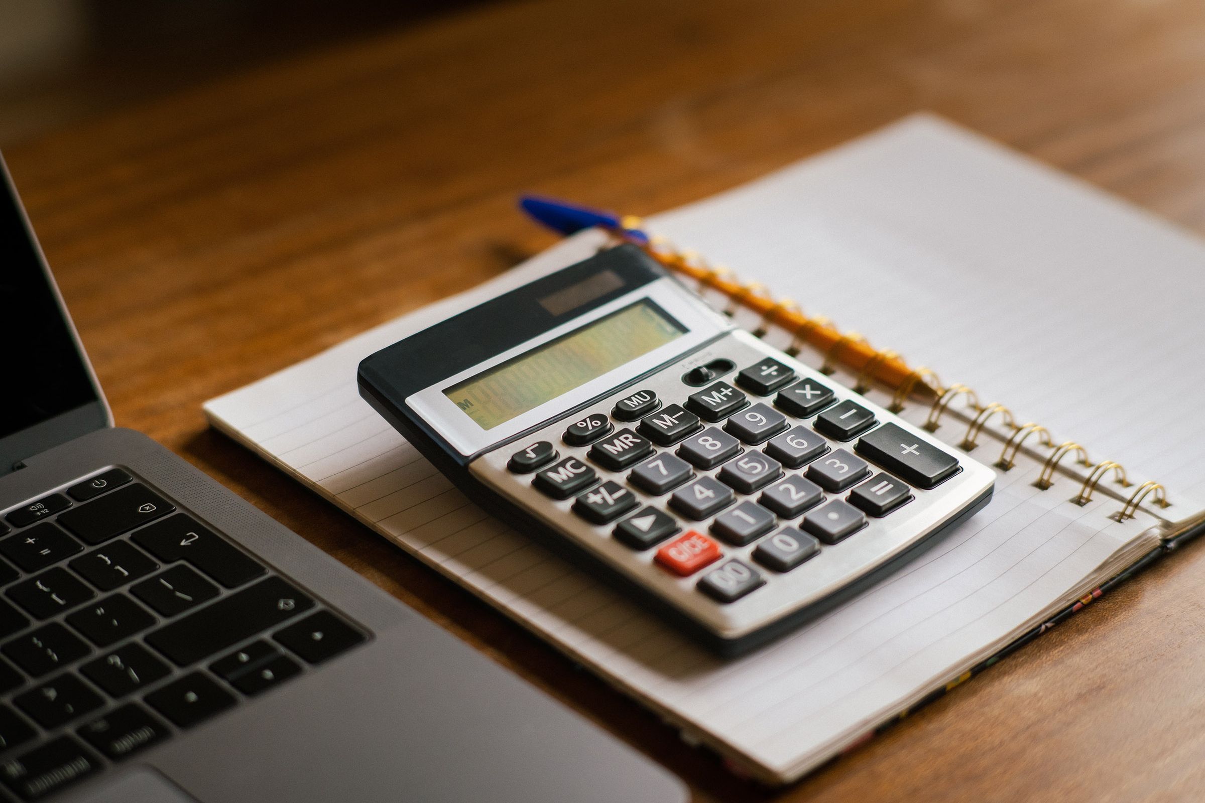 Calculadora sobre um caderno aberto, ao lado de um notebook, em uma mesa de madeira